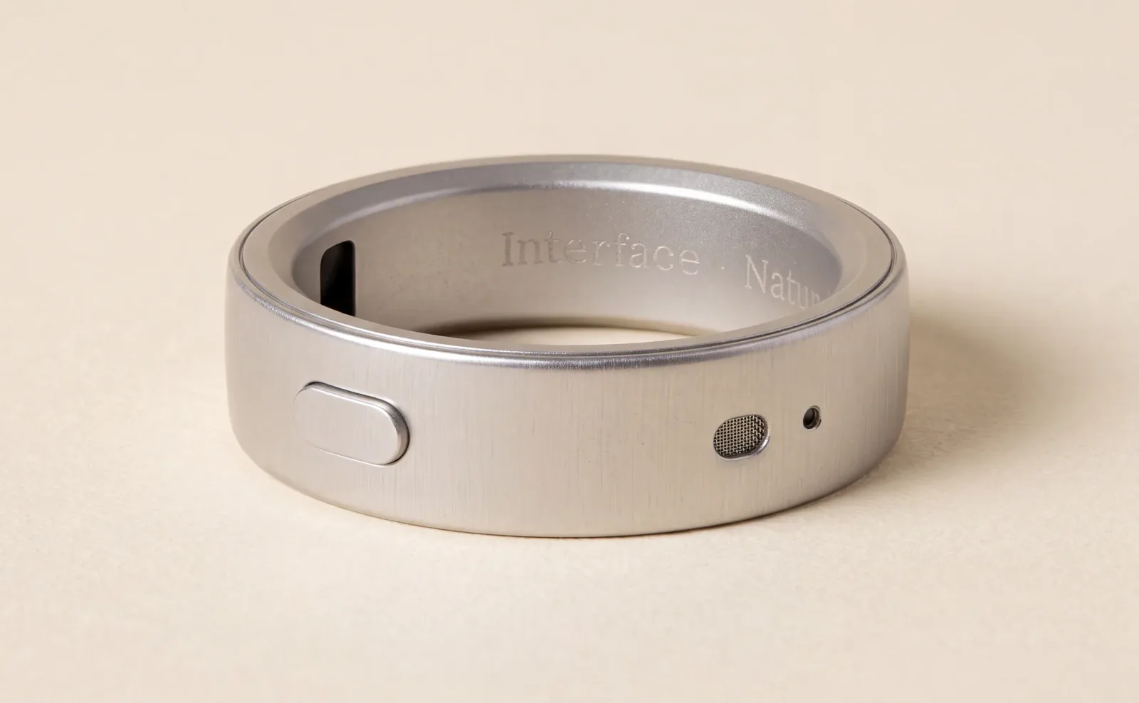 A silver Natura Interface smart ring with a physical button and microphone grille on a beige background.