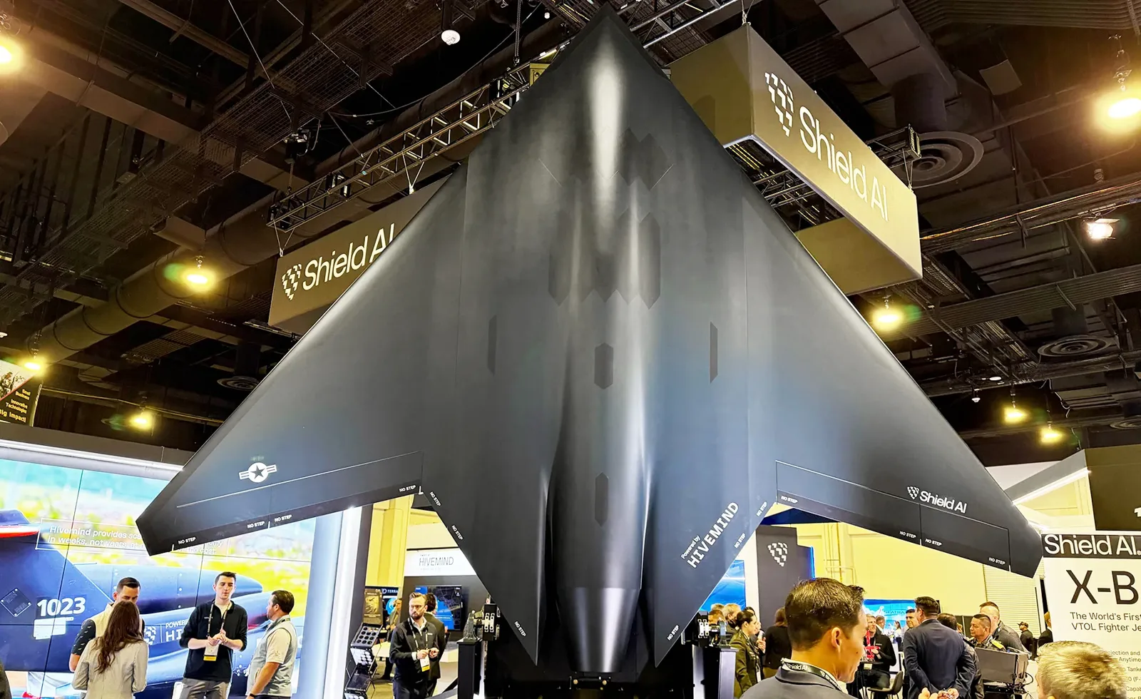 A large black delta-wing Shield AI X-BAT fighter drone displayed vertically at an exhibition booth with people looking at it.